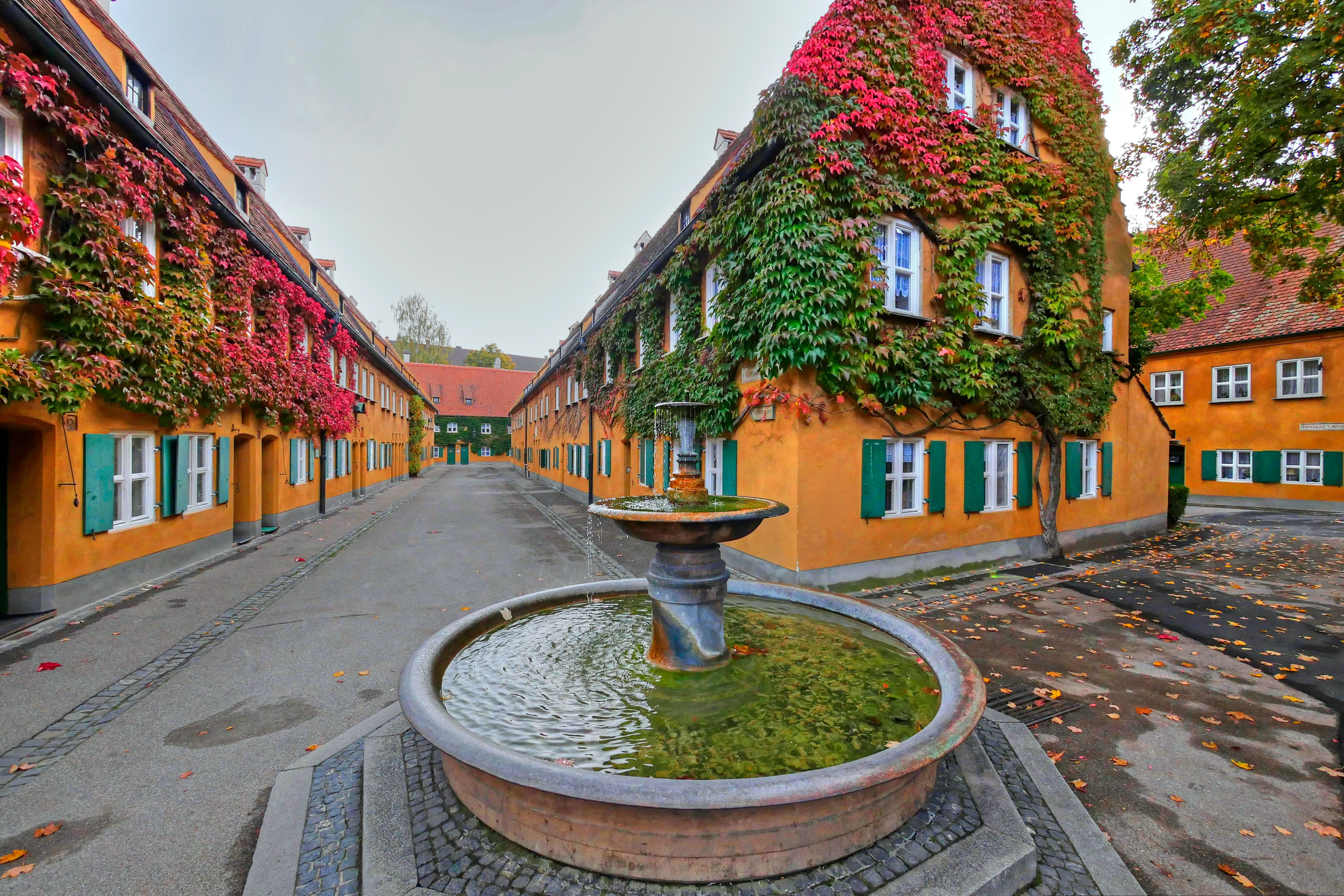 Augsburg Fuggerei, orangene Häuser mit Kletterpflanzen und davor ein Brunnen