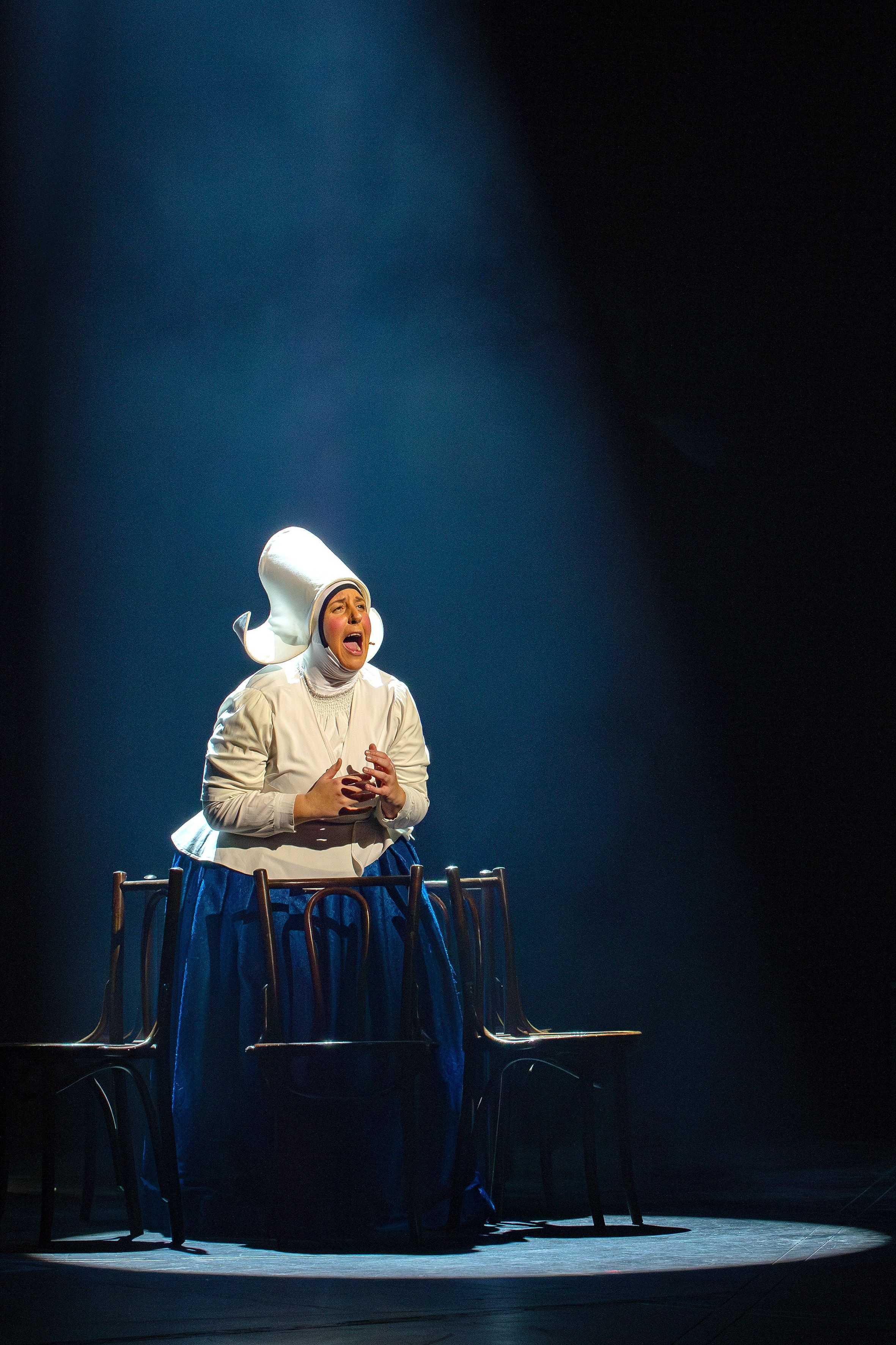Darstellerin in der Rolle der Amme singt auf der Bühne, umringt von drei Stühlen