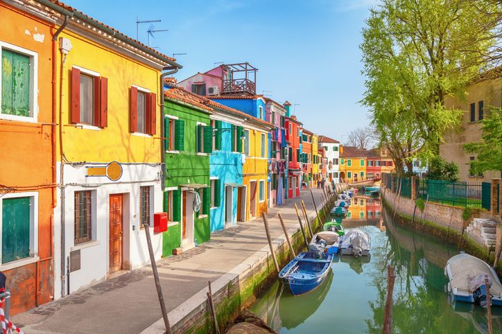 Farbenfrohe Häuserfassaden in Venedig säumen eine ruhige Wasserstraße. Die Gebäude in Gelb, Rot und Ocker spiegeln sich im Wasser, kleine Balkone, Fensterläden und Details verleihen der Szene mediterranen Charme.