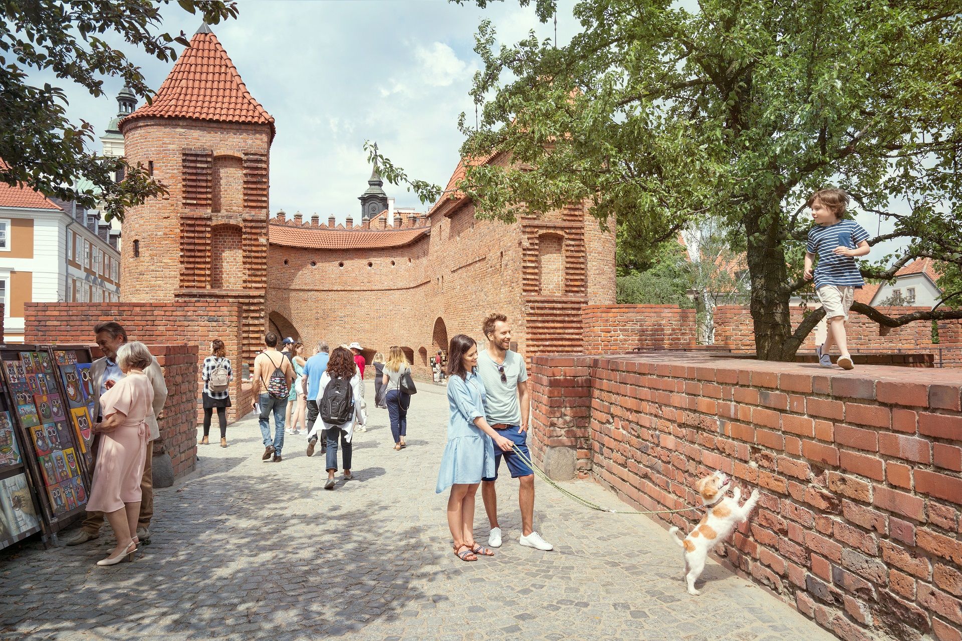Ein Paar spaziert mit einem Hund entlang des Barbakans in Warschau, einer historischen Stadtbefestigung aus rotem Backstein.