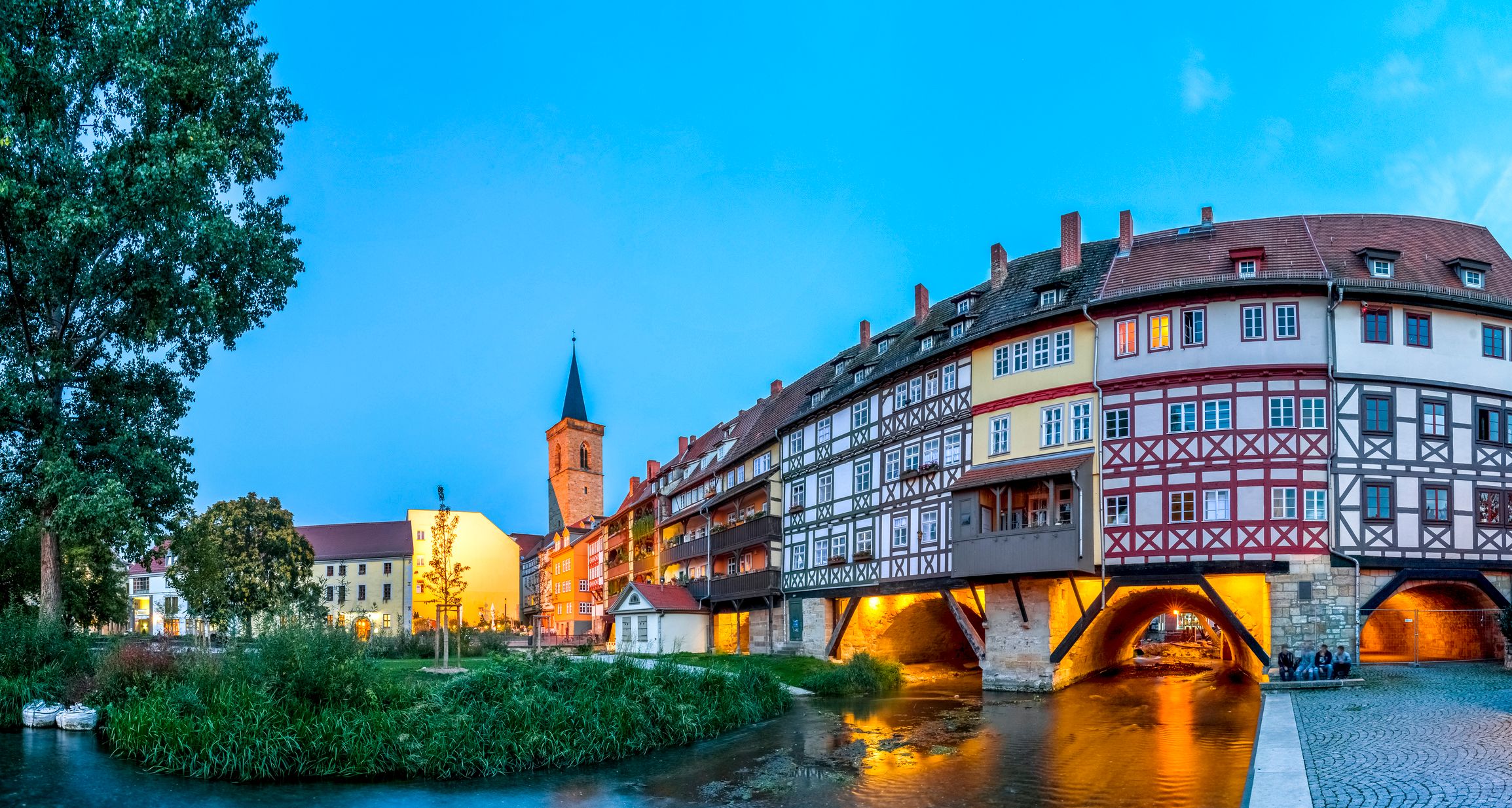 Weitwinkelaufnahme der Krämerbrücke in Erfurt bei Nacht, mit beleuchteten Fachwerkhäusern auf der Brücke und Spiegelung im Wasser der Gera.