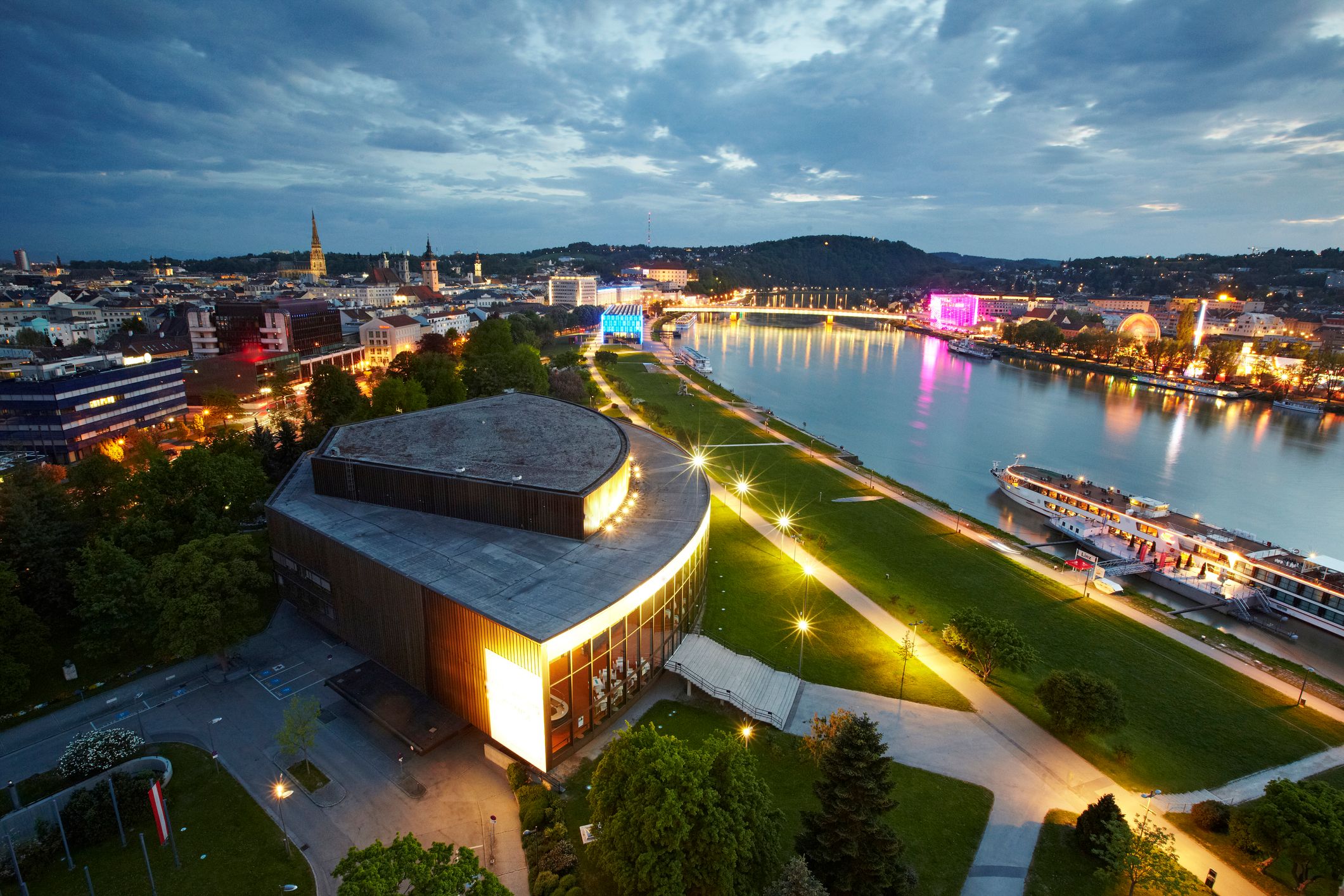 Die Donaulände in Linz bei Nacht – stimmungsvoll beleuchtete Promenade am Donauufer mit glitzernden Lichtern, die sich im Wasser spiegeln, und moderner Architektur im Hintergrund.