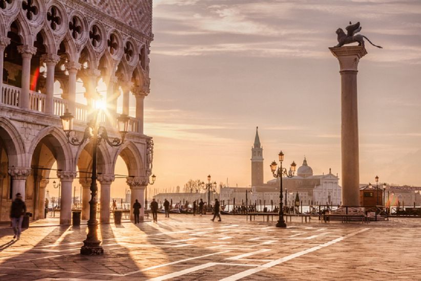 Der Markusplatz in Venedig im warmen Licht der untergehenden Sonne. Die Fassaden des Dogenpalasts und der Markusbasilika leuchten in goldenen Tönen, lange Schatten fallen über das Pflaster, während Menschen über den Platz flanieren.