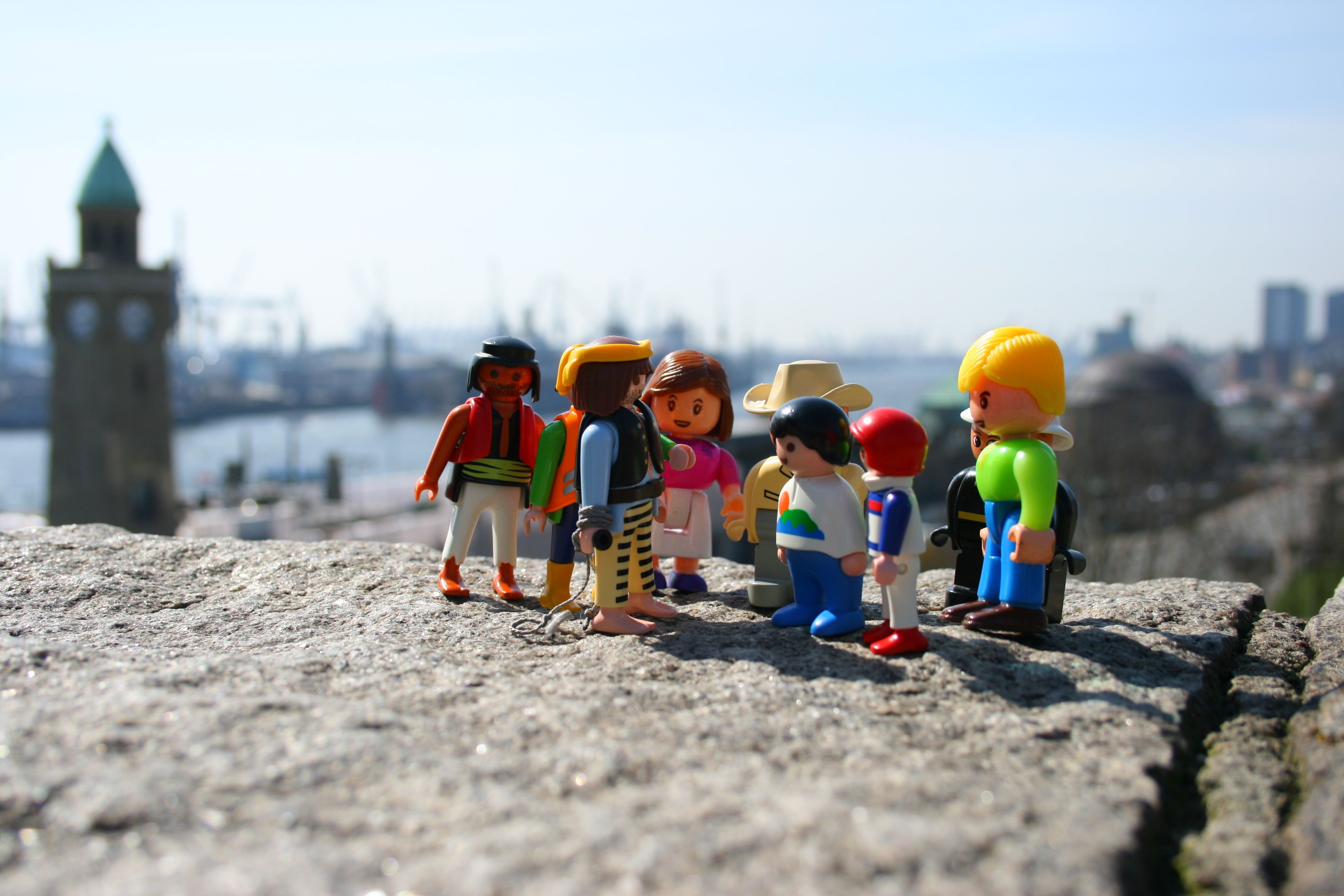 Mehrere Playmobil-Spielfiguren stehen im Vordergrund vor der Kulisse des Hamburger Hafens mit den markanten Kränen, Containerschiffen und den Landungsbrücken bei Tageslicht.