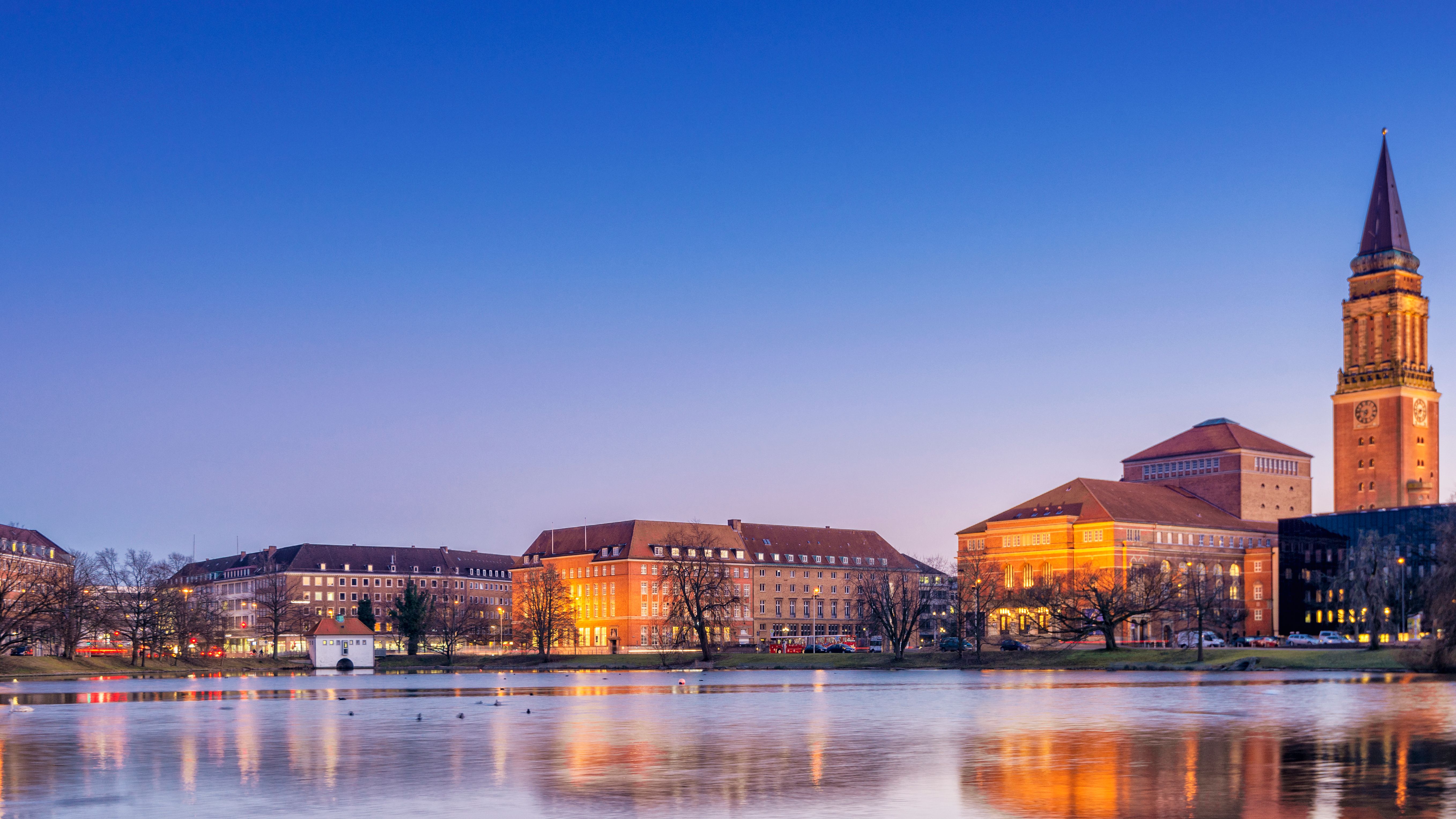 Abendlicher Blick über die ruhige Ostsee auf die Stadt Kiel mit beleuchteter Uferlinie und sanftem Licht am Horizont.