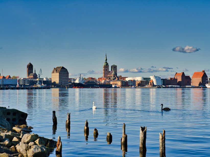 Panoramablick auf das Ufer von Stralsund mit historischen Giebelhäusern, Hafenanlagen und ruhigem Wasser im Vordergrund.