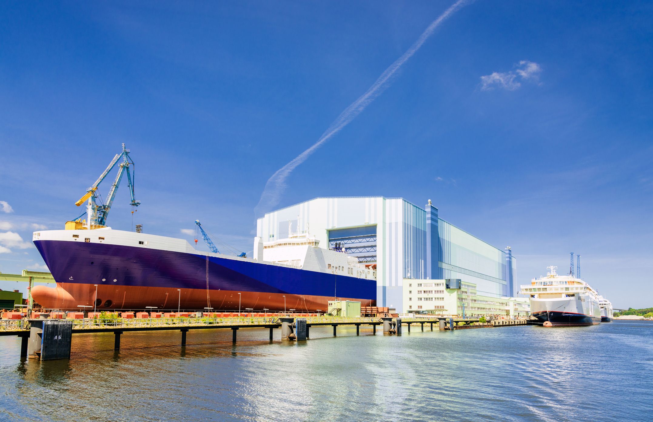 Große Werfthalle in Stralsund mit einem teils fertiggestellten Schiff im Inneren, umgeben von industrieller Infrastruktur.