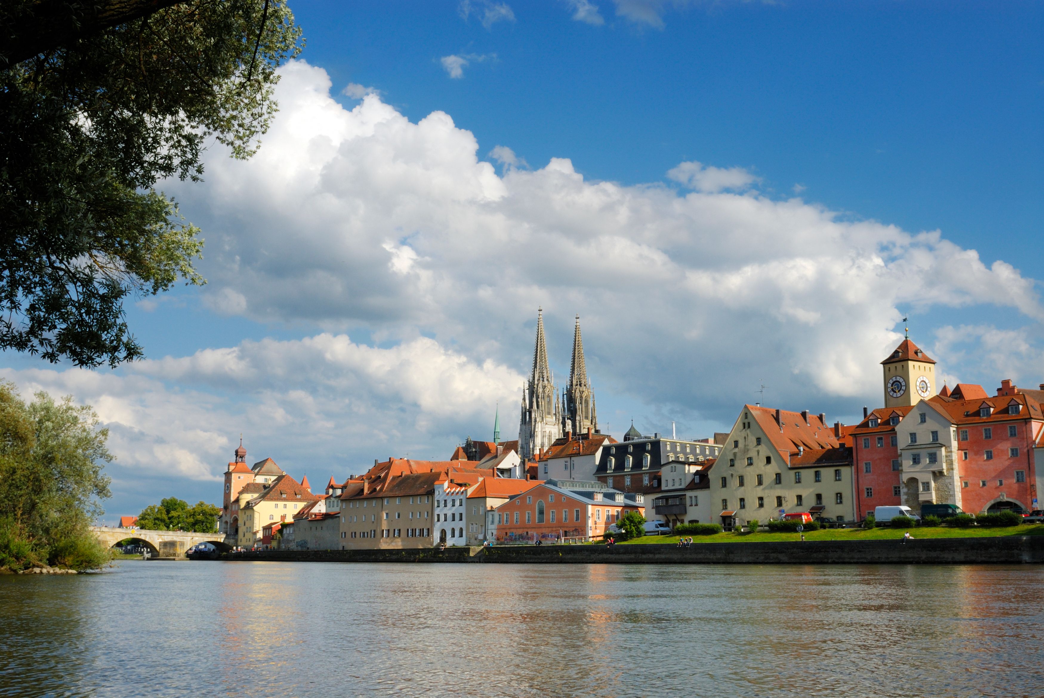 Panoramablick vom Ufer der Donau auf die Steinerne Brücke und die Altstadt von Regensburg mit historischen Gebäuden und Kirchtürmen.