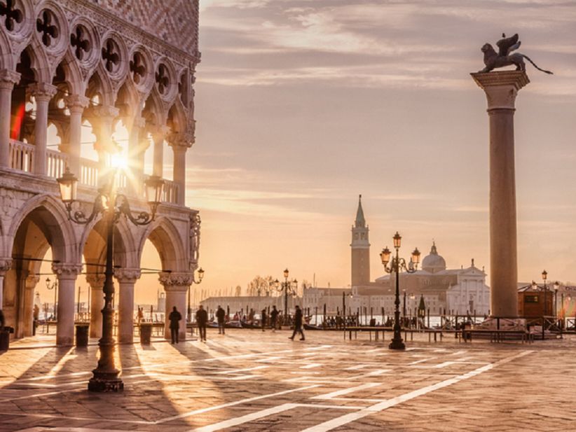 Der Markusplatz in Venedig im warmen Licht der untergehenden Sonne. Die Fassaden des Dogenpalasts und der Markusbasilika leuchten in goldenen Tönen, lange Schatten fallen über das Pflaster, während Menschen über den Platz flanieren.