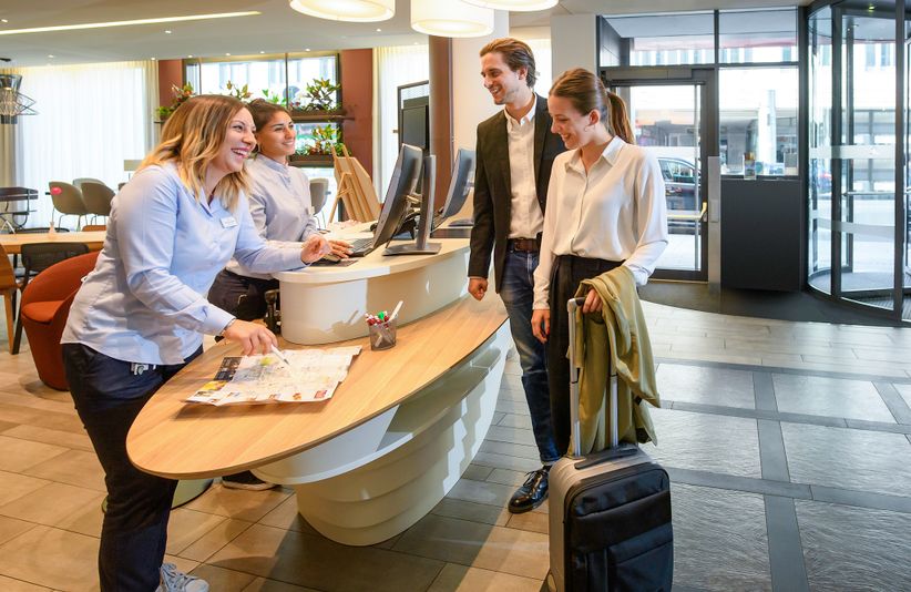 Zwei Besucher stehen an der Rezeption des Adagio Aparthotel in München mit zwei Rezeptionistinnen