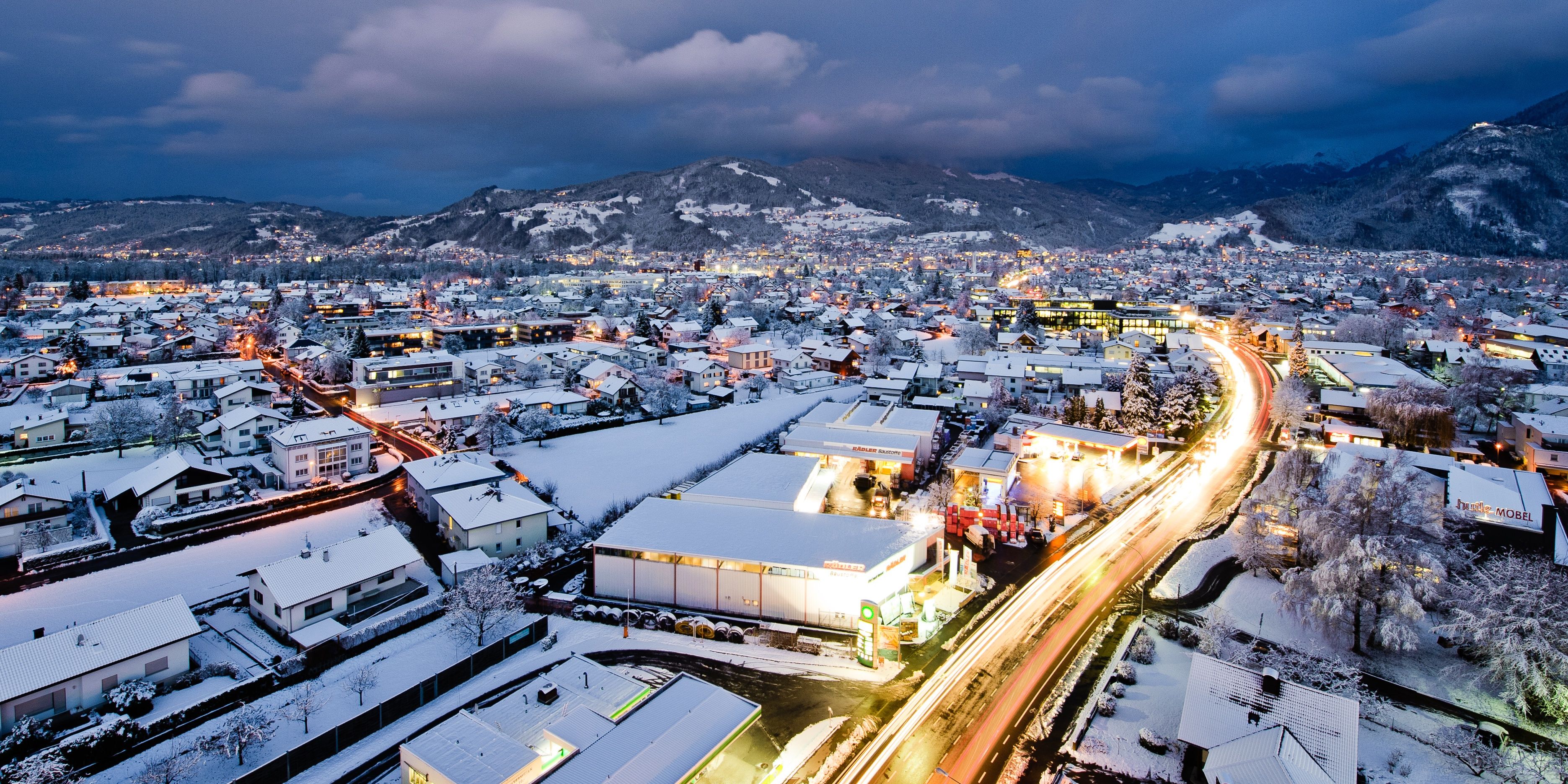 Panoramablick auf das verschneite Dornbirn mit leuchtenden Straßen und dahinterliegende schneebedeckte Berge 