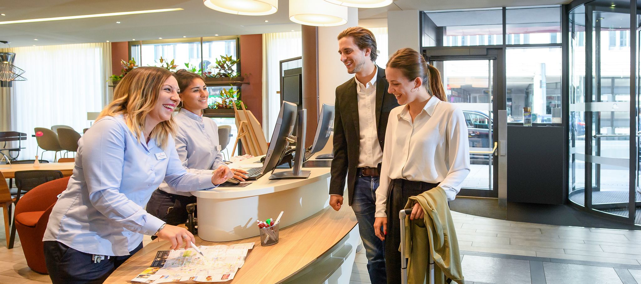 Zwei Besucher stehen an der Rezeption des Adagio Aparthotel in München mit zwei Rezeptionistinnen