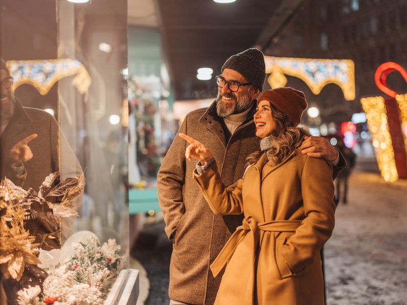 Ein warm eingepacktes Paar schaut sich ein leuchtendes Schaufenster an, im Hintergrund ist Weihnachtsdeko und Schnee