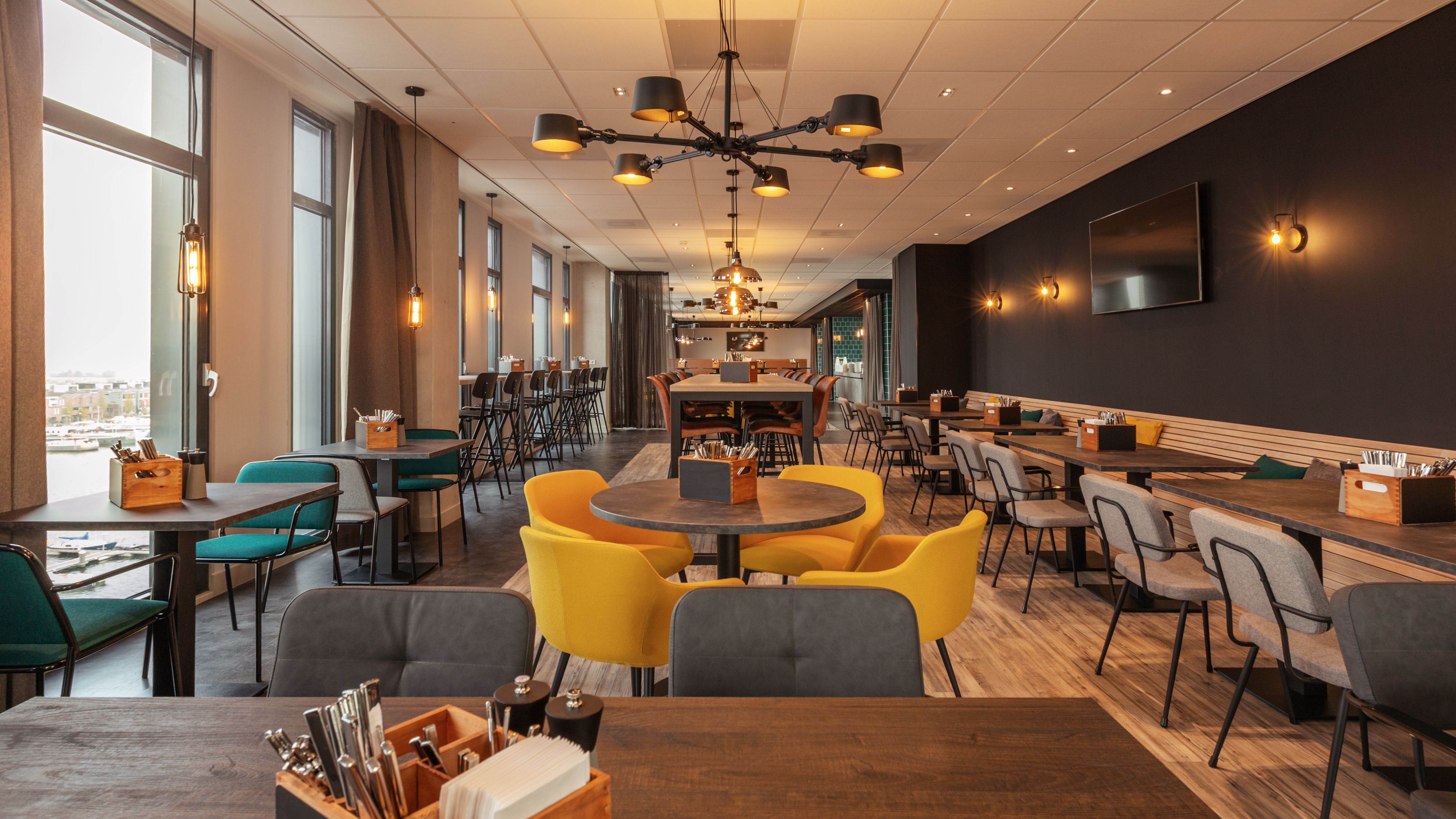 Moderner Frühstücksbereich im Hotel the niu Fender Amsterdam mit industriellem Design, hohen Decken, stylischen Pendelleuchten und einer Kombination aus rustikalen Holztischen und Metallelementen im Hafen-Stil.