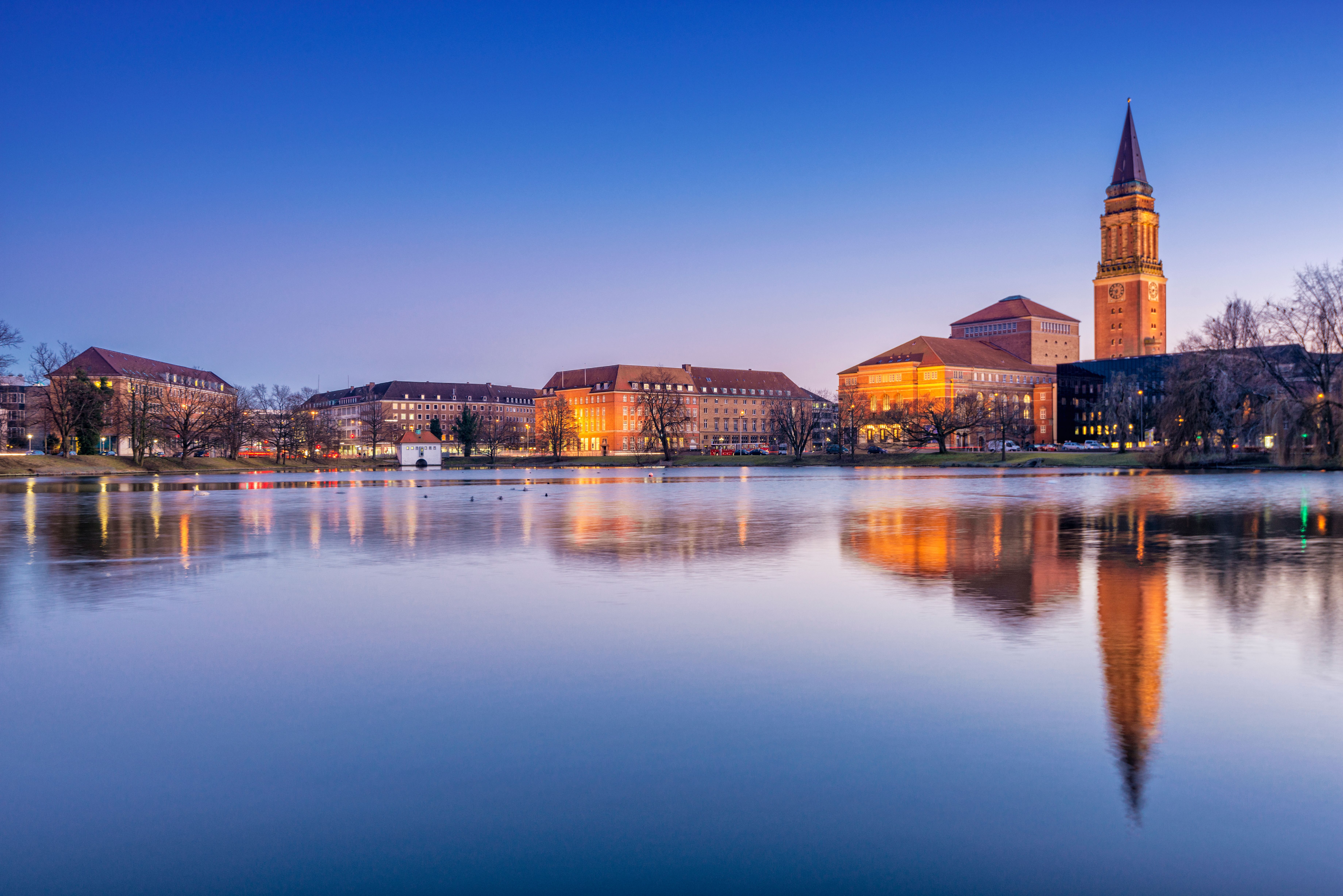 Abendlicher Blick über die ruhige Ostsee auf die Stadt Kiel mit beleuchteter Uferlinie und sanftem Licht am Horizont.