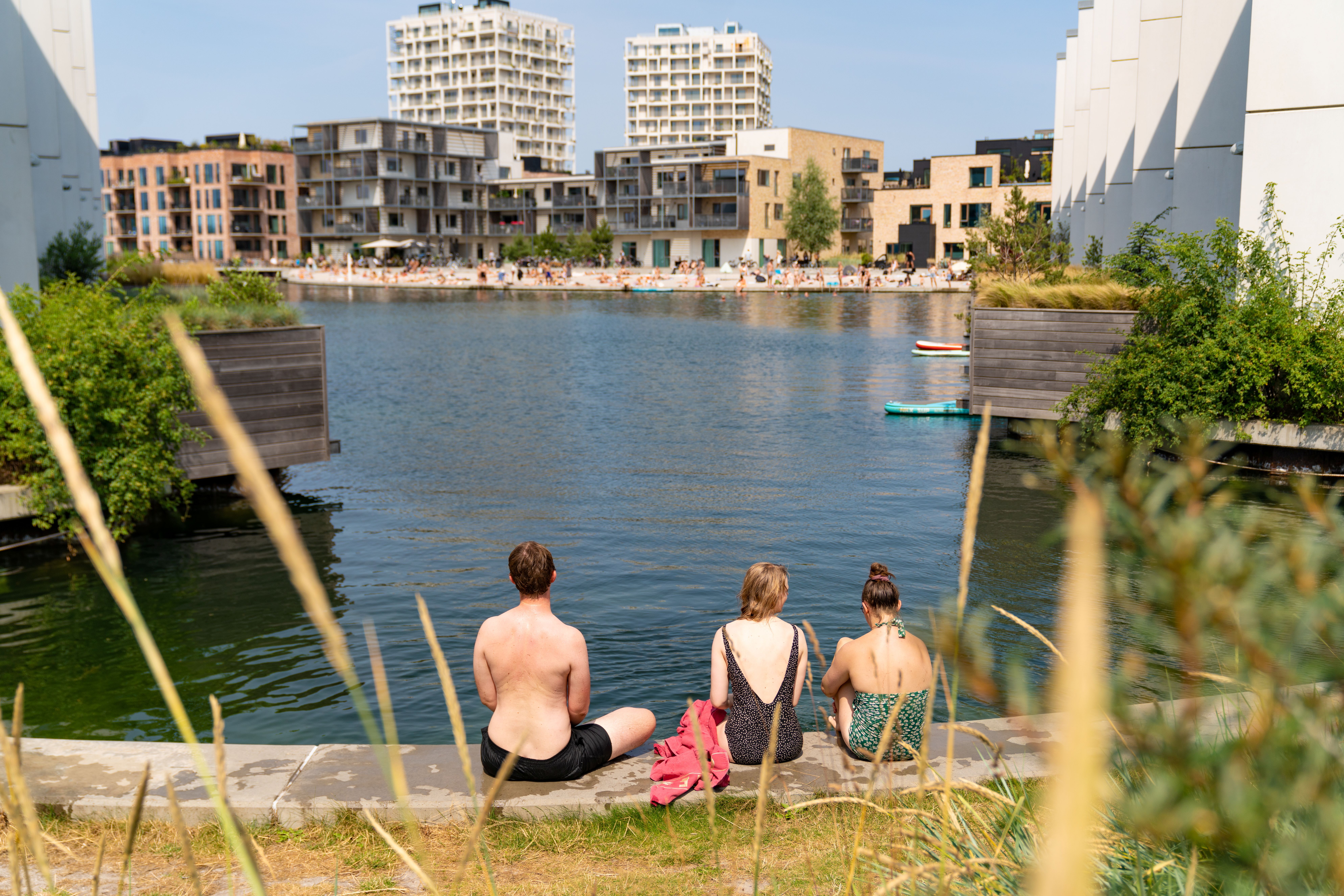 drei Menschen in Badebekleidung sitzen am Rand eines Hafenbeckens in Kopenhagen im Sommer