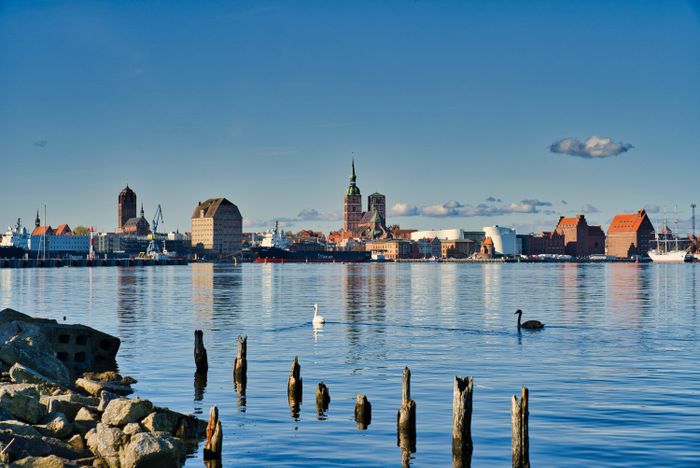 Panoramablick auf das Ufer von Stralsund mit historischen Giebelhäusern, Hafenanlagen und ruhigem Wasser im Vordergrund.