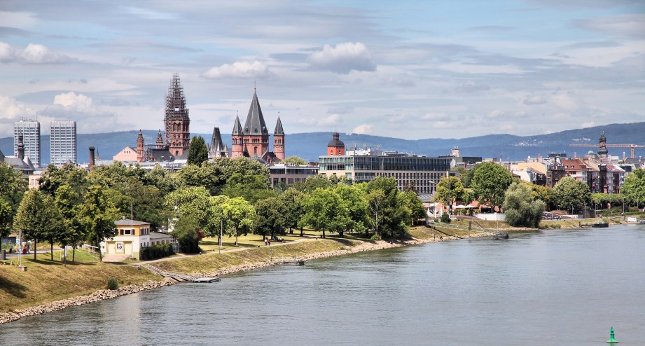 Mainz, Deutschland, Stadt in Rheinland-Pfalz. Die City-Skyline zwischen Bäumen zeichnet sich unter einem Himmel mit Wolken ab, Hügellandschaft im Hintergrund - BAHNHIT.DE, © getty, Foto: tupungato