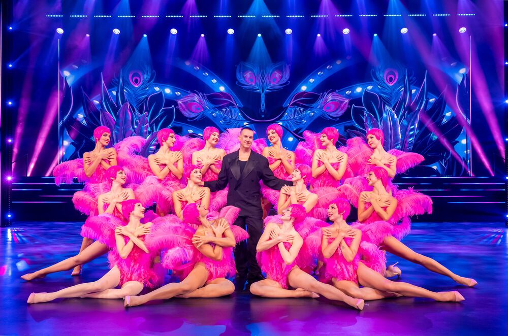 Viele Damen in pinken Federkostümen stehen um einen Anzugträger auf der Bühne