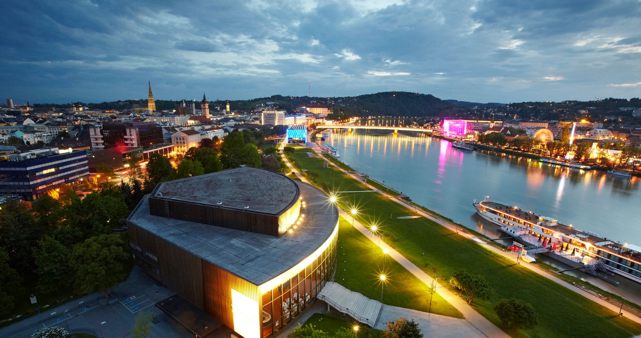 Die Donaulände in Linz bei Nacht – stimmungsvoll beleuchtete Promenade am Donauufer mit glitzernden Lichtern, die sich im Wasser spiegeln, und moderner Architektur im Hintergrund.