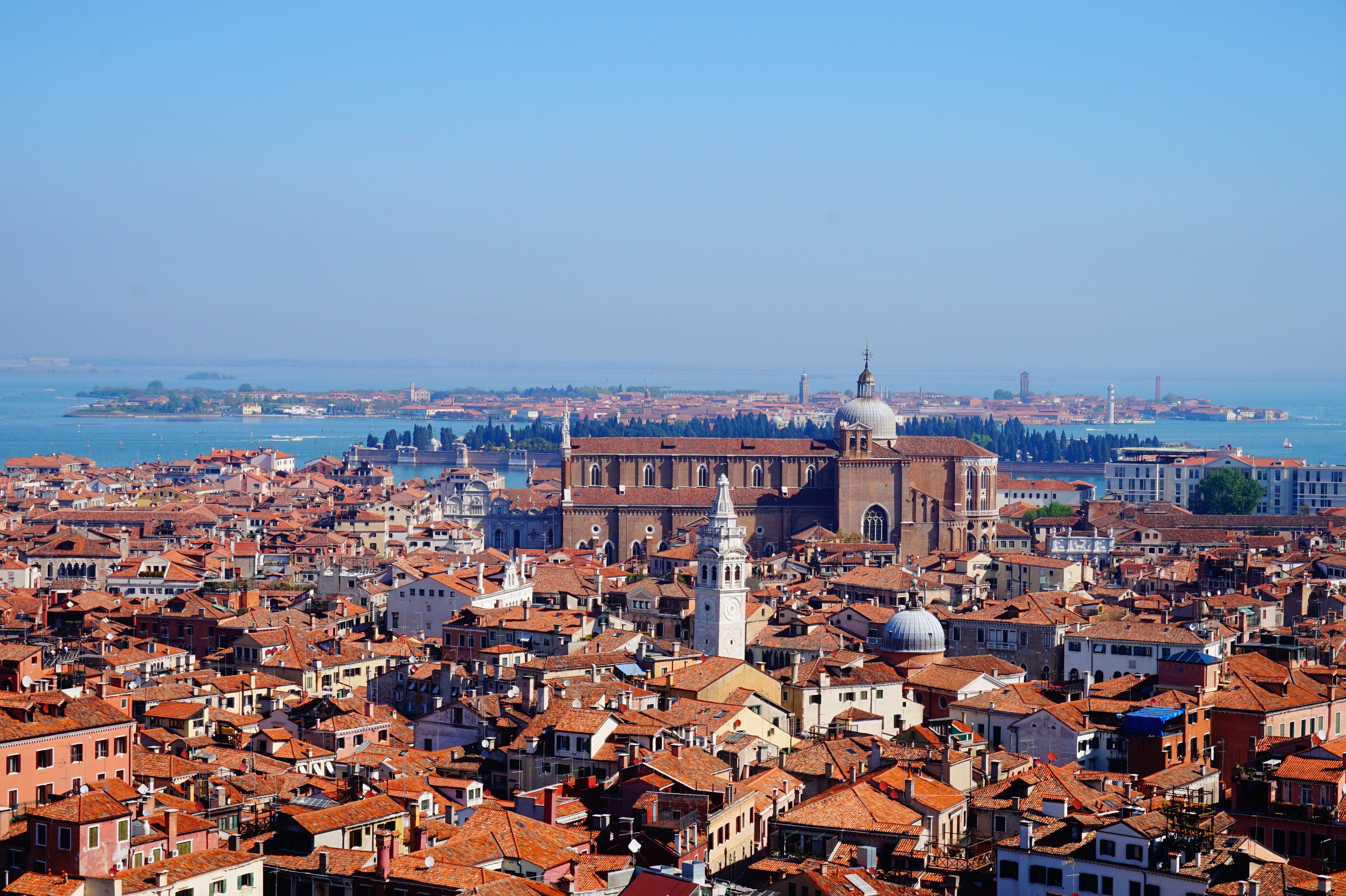 Blick von oben auf Venedig: Ein dichtes Meer aus roten Ziegeldächern zieht sich durch enge Gassen und Kanäle. Im Hintergrund glitzert die Lagune, dazwischen ragen markante Kuppeln und der Campanile empor – ein Panorama der historischen Altstadt aus der Vogelperspektive.