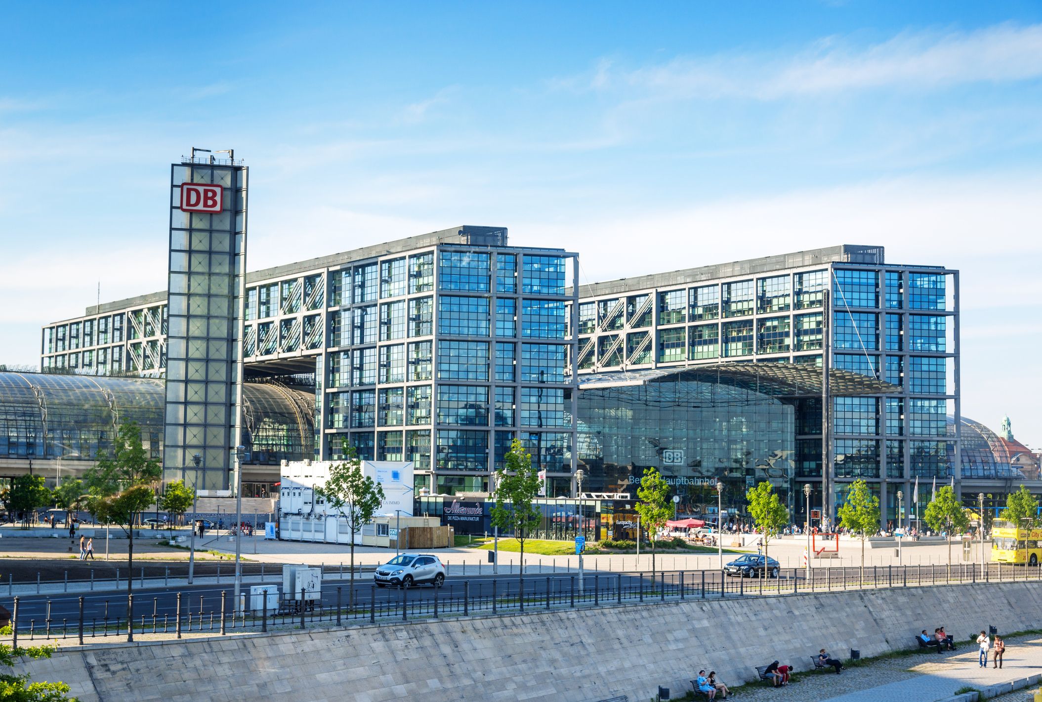 Außenansicht des Berliner Hauptbahnhofs bei Tag: Die markante Glasfassade und die geschwungene Dachkonstruktion spiegeln den Himmel wider, davor befinden sich der Bahnhofsvorplatz und Passanten.