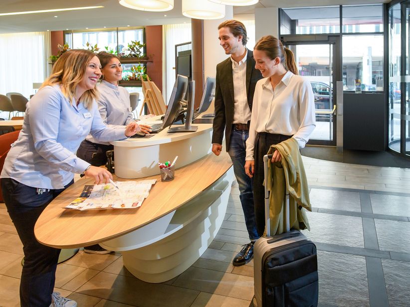 Zwei Besucher stehen an der Rezeption des Adagio Aparthotel in München mit zwei Rezeptionistinnen
