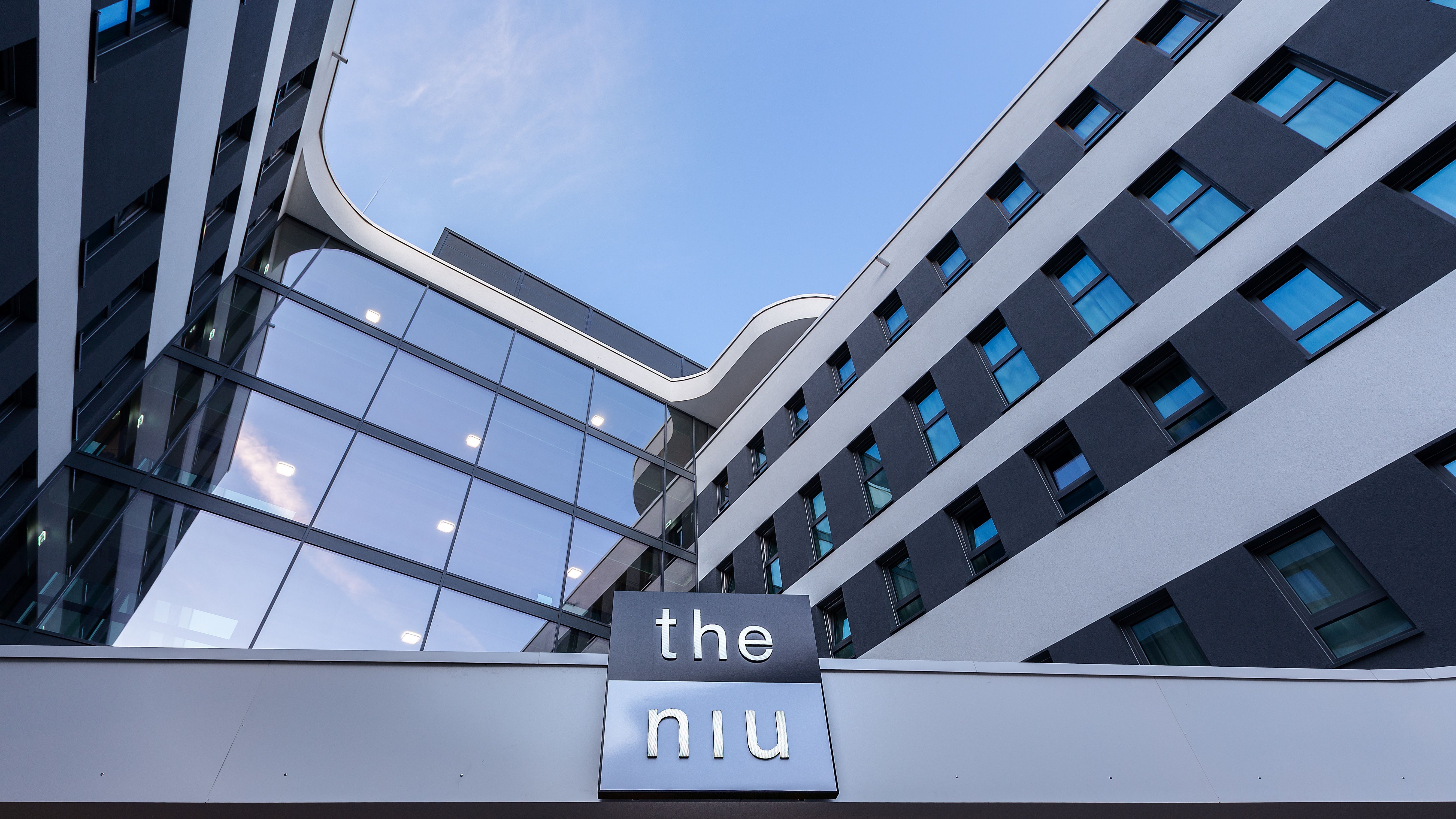 Moderne Außenansicht des Hotels the niu Mesh in Stuttgart mit markanter, gemusterter Fassade, großen Fensterfronten und dem sichtbaren Hotellogo vor einem klaren Himmel.