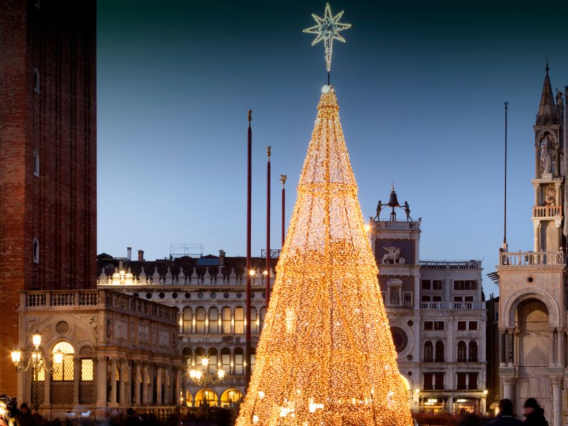 Der festlich erleuchtete Weihnachtsbaum auf dem Markusplatz in Venedig bei Nacht. Der Baum ist mit Lichtern geschmückt und trägt einen leuchtenden Stern auf der Spitze, im Hintergrund sind historische Fassaden und weihnachtliche Stimmung zu erkennen.